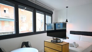 Hotelkamer met een bed, een televisie op een houten dressoir, een kleine tafel en grote ramen met uitzicht op gebouwen.