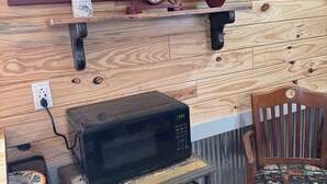 Interior de madera con un reloj, un microondas sobre una mesita y una silla con cojín estampado.
