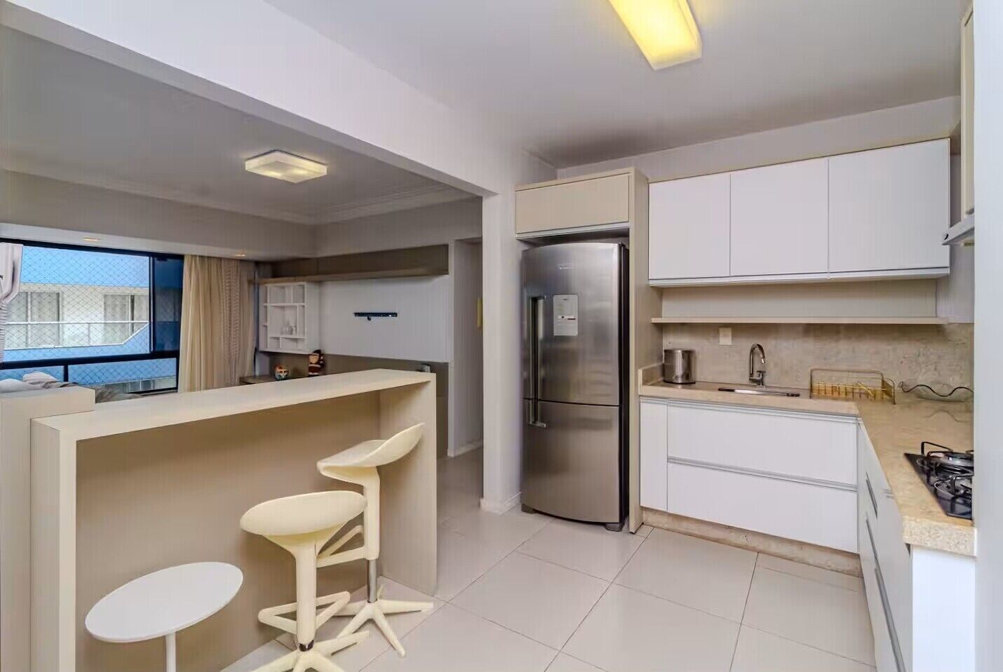 A modern kitchen with white cabinetry, a stainless steel refrigerator, and a bar area with stools.