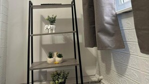 A corner of a room with a black metal shelf unit holding decorative items, a window with a curtain, and a textured white wall. - Comfortable Stay with Free Parking (Moor Row)