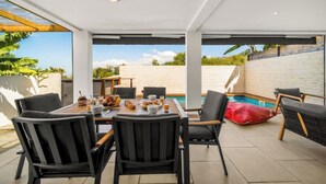 A modern outdoor dining area with a pool view, featuring a wooden table and black chairs. - Villa Rêve Austral (Saint-Pierre)