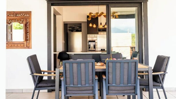 A dining area with a table and four chairs, a wooden-framed mirror on the wall, and a view of the kitchen and outdoors through a glass door. - Villa Rêve Austral (Saint-Pierre)