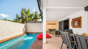 A swimming pool with a wooden deck, a dining area with chairs and a table, and a view of a garden with trees. - Villa Rêve Austral (Saint-Pierre)