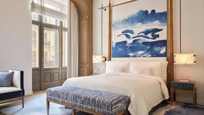 Un dormitorio con una cama grande, un banco, una mesita de noche, una obra de arte en la pared y vistas a través de una ventana.