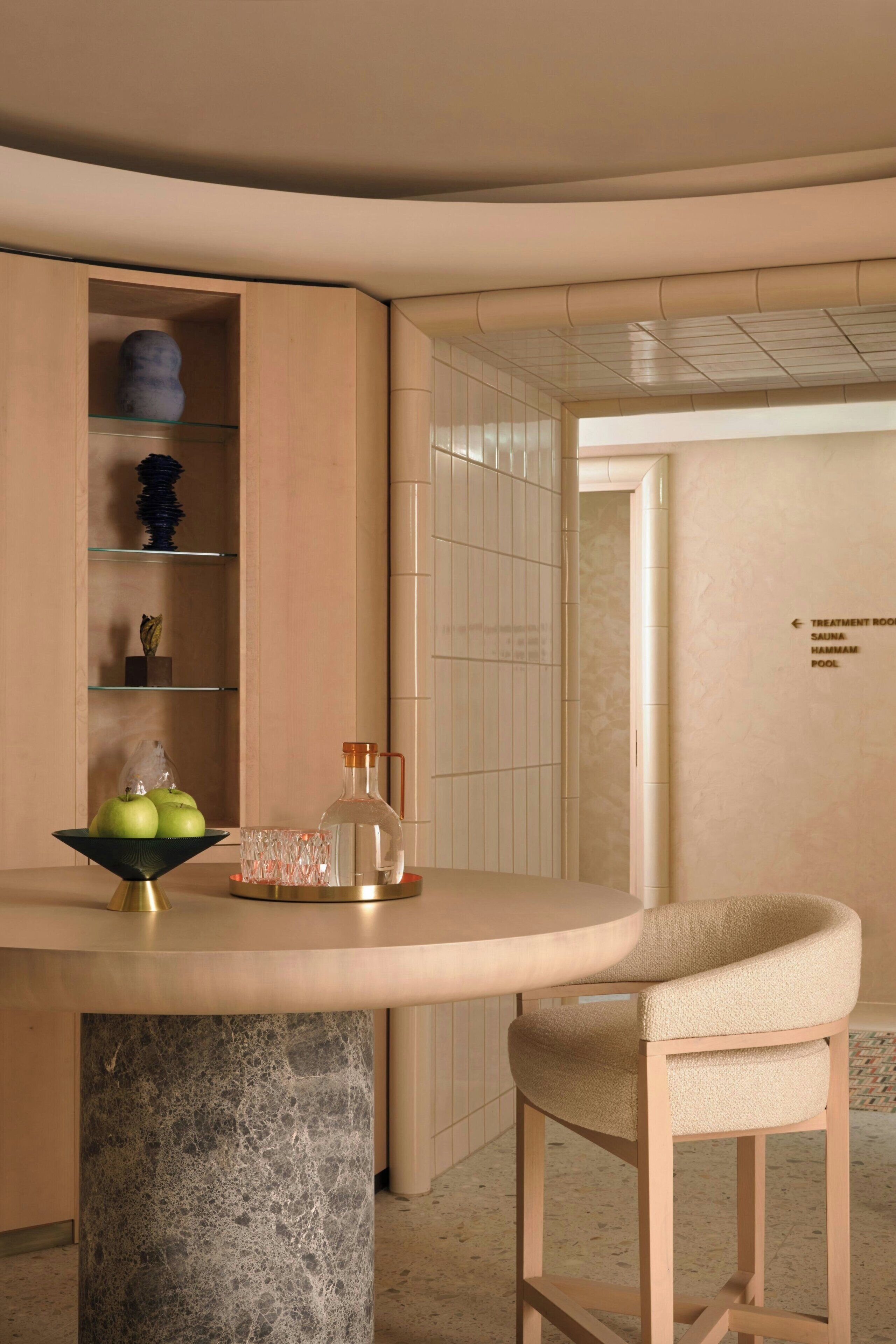 Une salle de bain moderne avec une colonne en marbre, une table ronde surmontée d’une carafe en verre et une chaise. On y trouve des étagères ornées d’objets décoratifs, ainsi qu’un panneau indiquant l’emplacement des salles de soins et de la piscine.