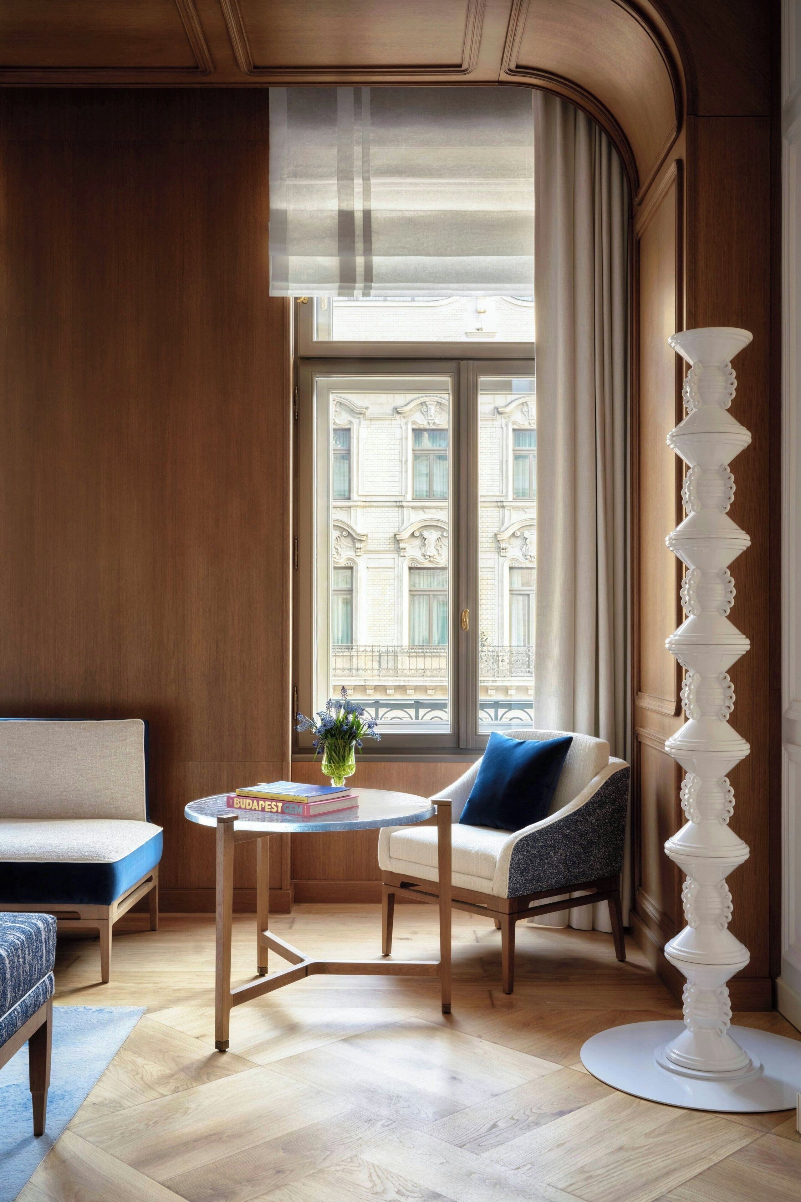 A room with a wooden paneled wall, a round table with a book, a blue chair, a white floor lamp, and a window with a view of buildings.