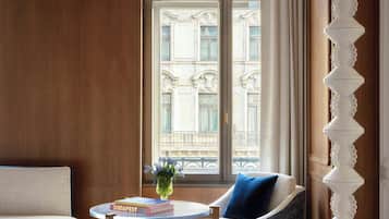 A room with a wooden paneled wall, a round table with a book, a blue chair, a white floor lamp, and a window with a view of buildings.