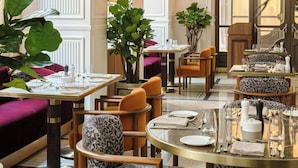 Comedor con mesas dispuestas para dos, sillas tapizadas y una planta grande en maceta.
