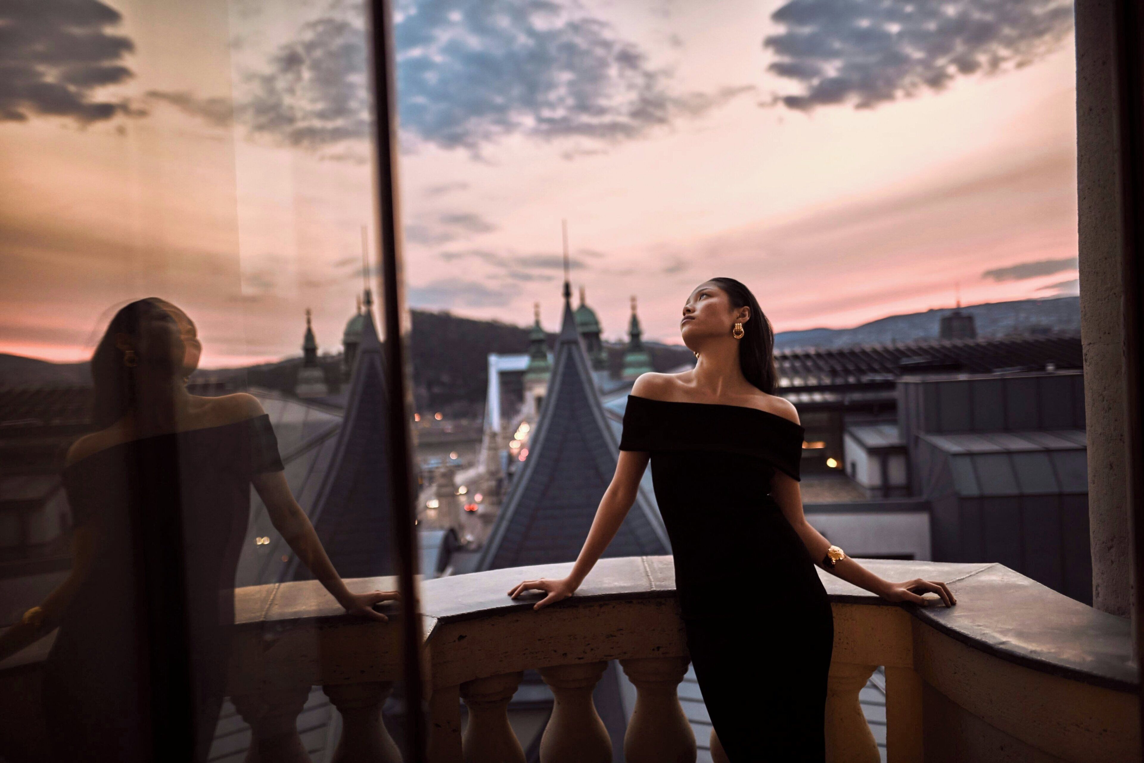 A person in a black dress standing by a window overlooking a cityscape with distinctive architecture.