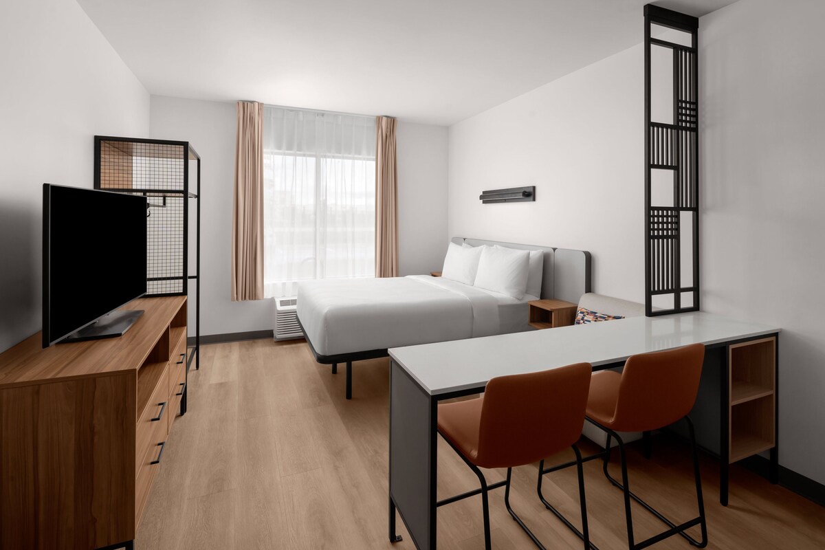A modern hotel room with a bed, desk, chairs, a TV, and a window with curtains.