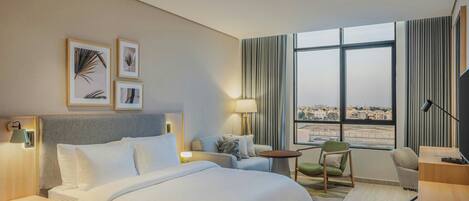 A hotel room with a large bed, a sofa, a desk, a TV, and a window with a city view.