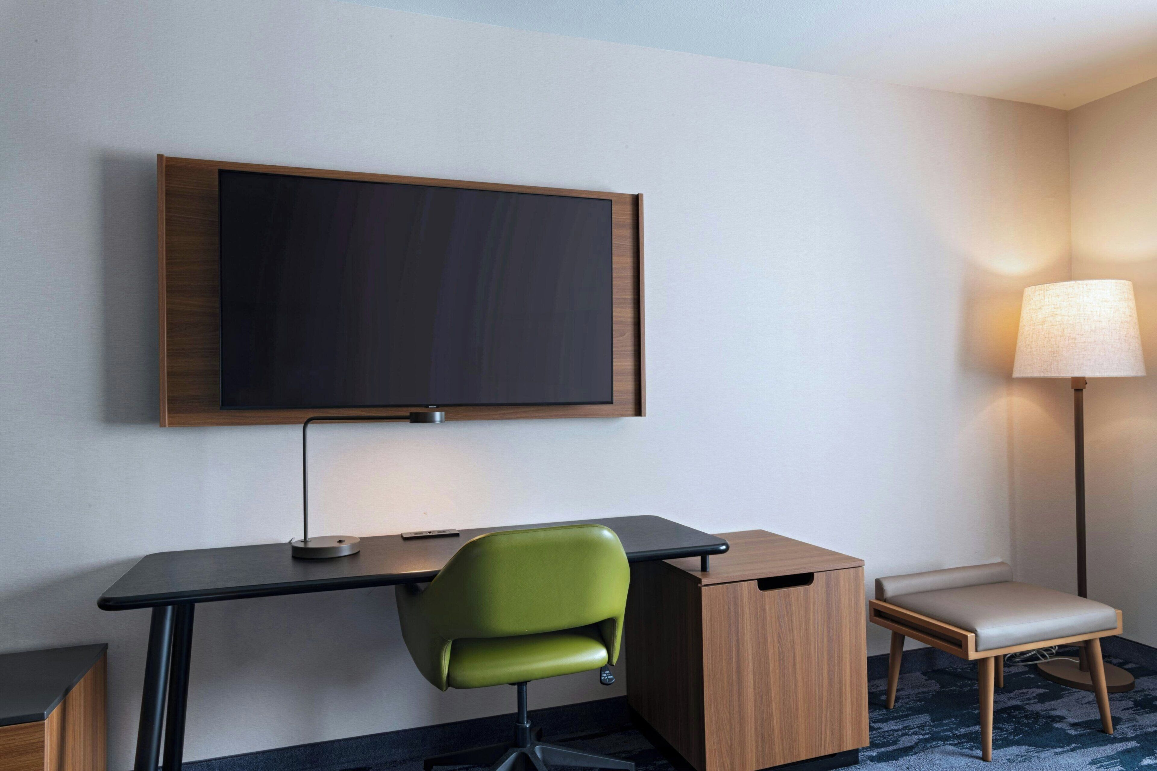 Quarto de hotel com televisão de tela plana na parede, escrivaninha com cadeira, mesa pequena e abajur.