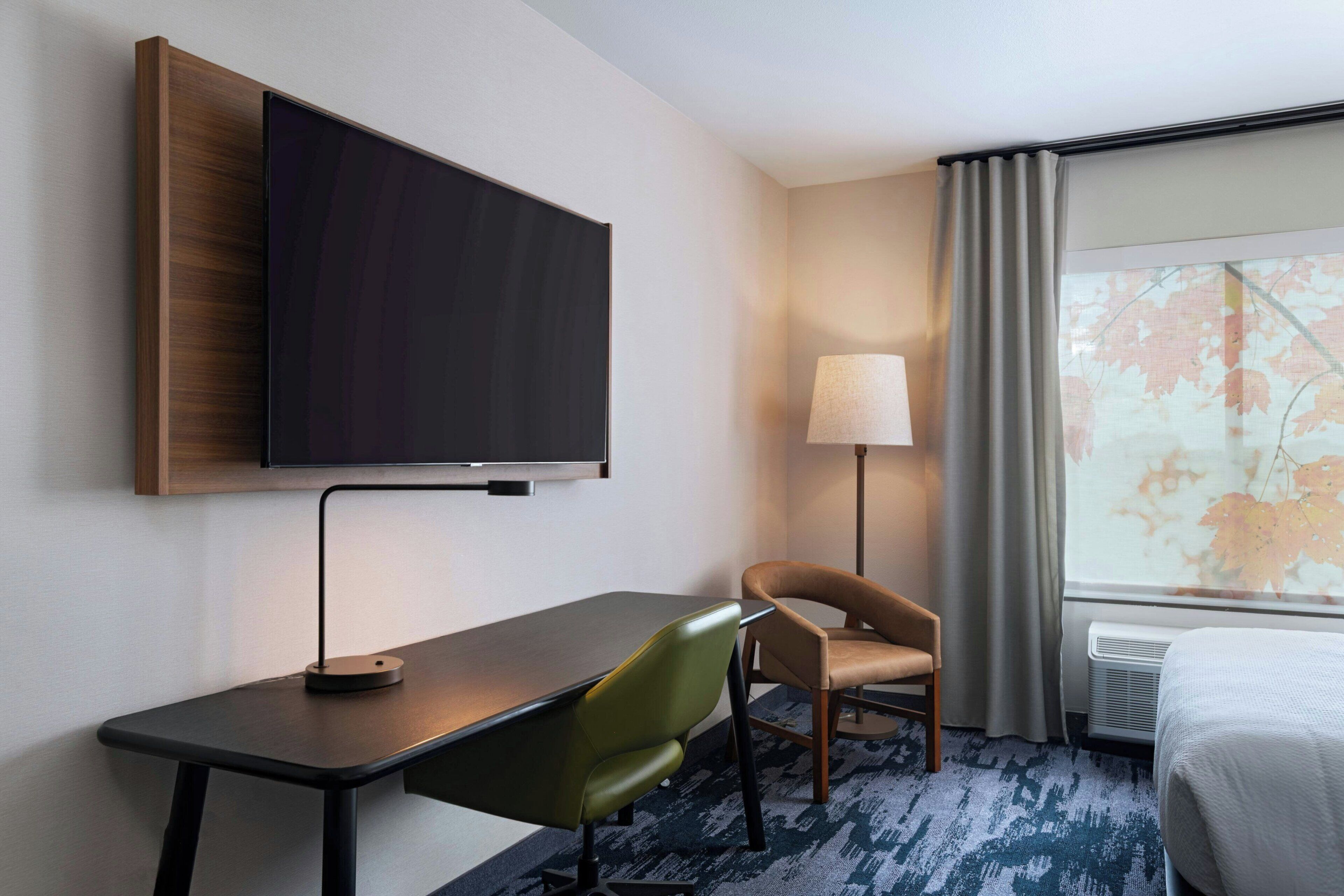 Habitación de hotel con televisión de pantalla plana montada en la pared, escritorio con silla, lámpara y cama.