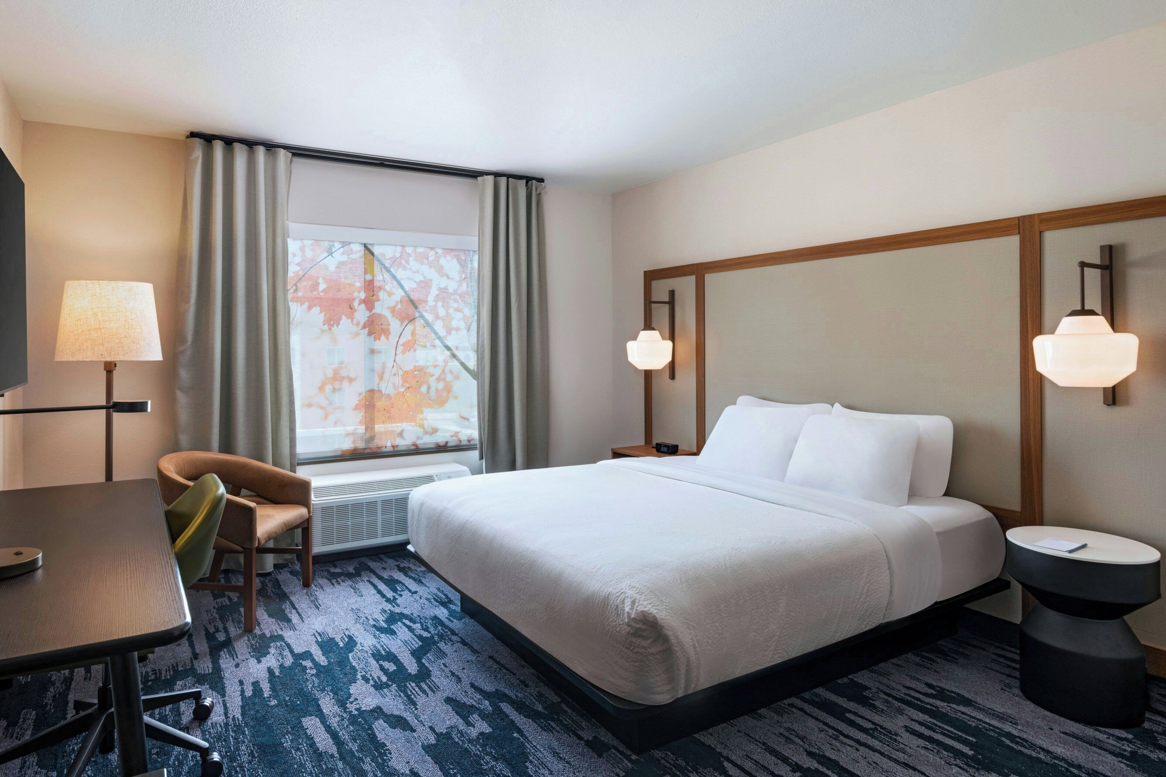A hotel room with a bed, a desk, a chair, a nightstand, and a lamp.