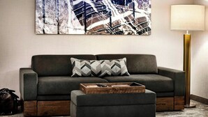 Una sala de estar moderna con un sofá gris, un puf a juego y una mesita de madera. Un cuadro abstracto grande adorna la pared, y una lámpara de pie está junto al sofá.