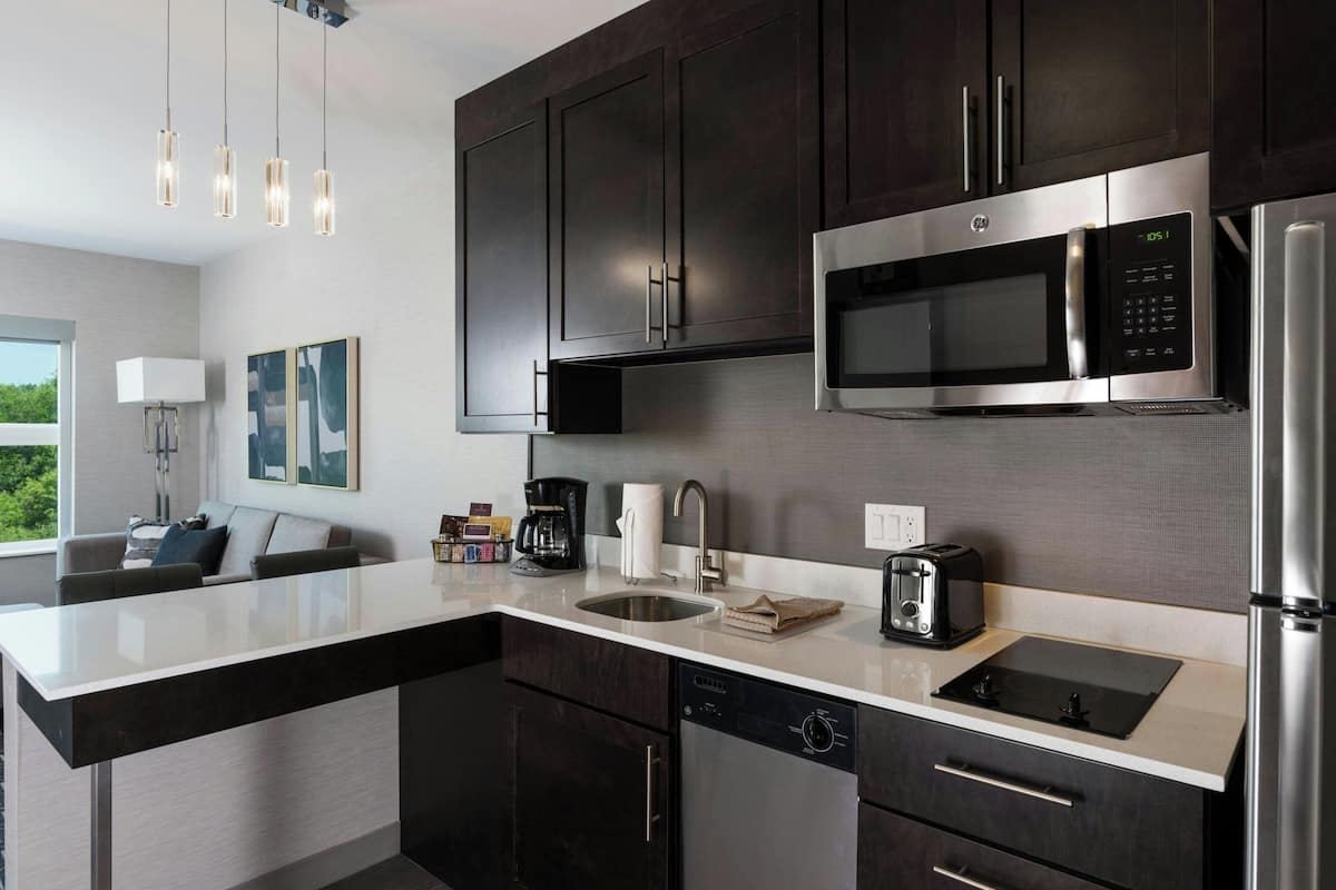 A modern kitchen with dark cabinets, stainless steel appliances, a large island with a sink, and a dining area with a sofa and coffee machine.