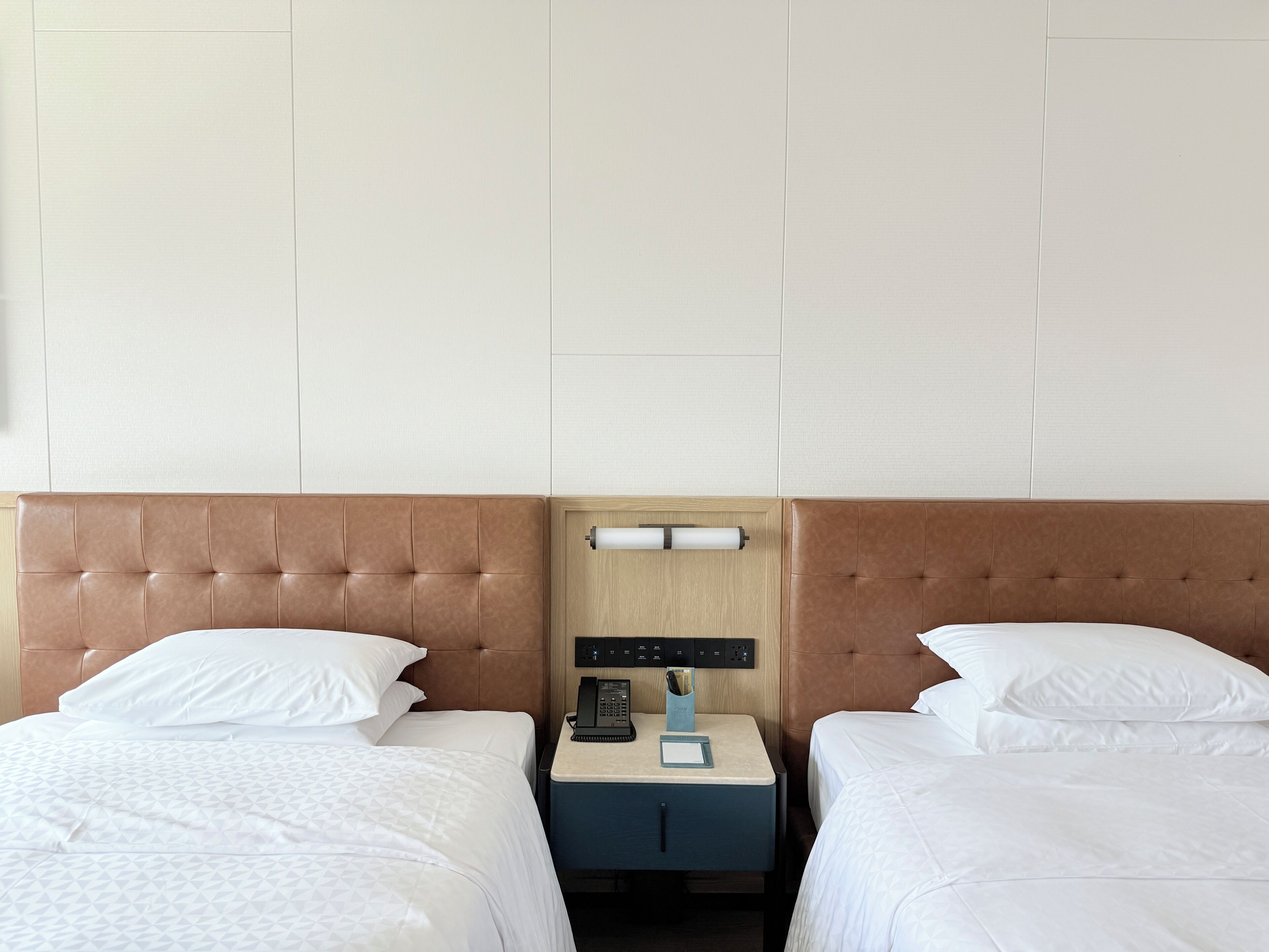 A hotel room with two beds, a nightstand with a phone and a lamp, and a wall-mounted control panel.