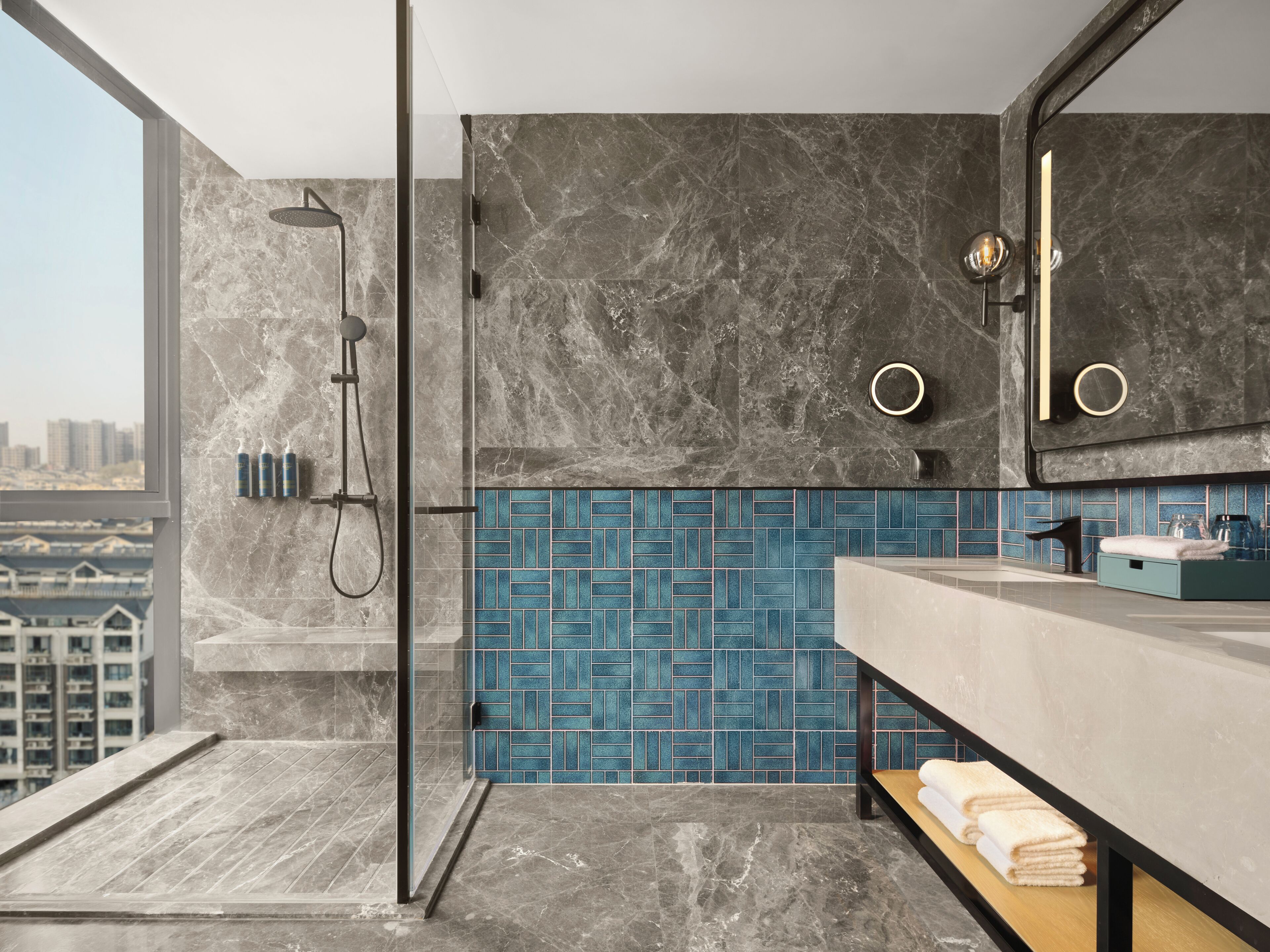 A modern bathroom with a glass-enclosed shower, a blue-tiled wall, a large mirror, and a wooden bench with towels.