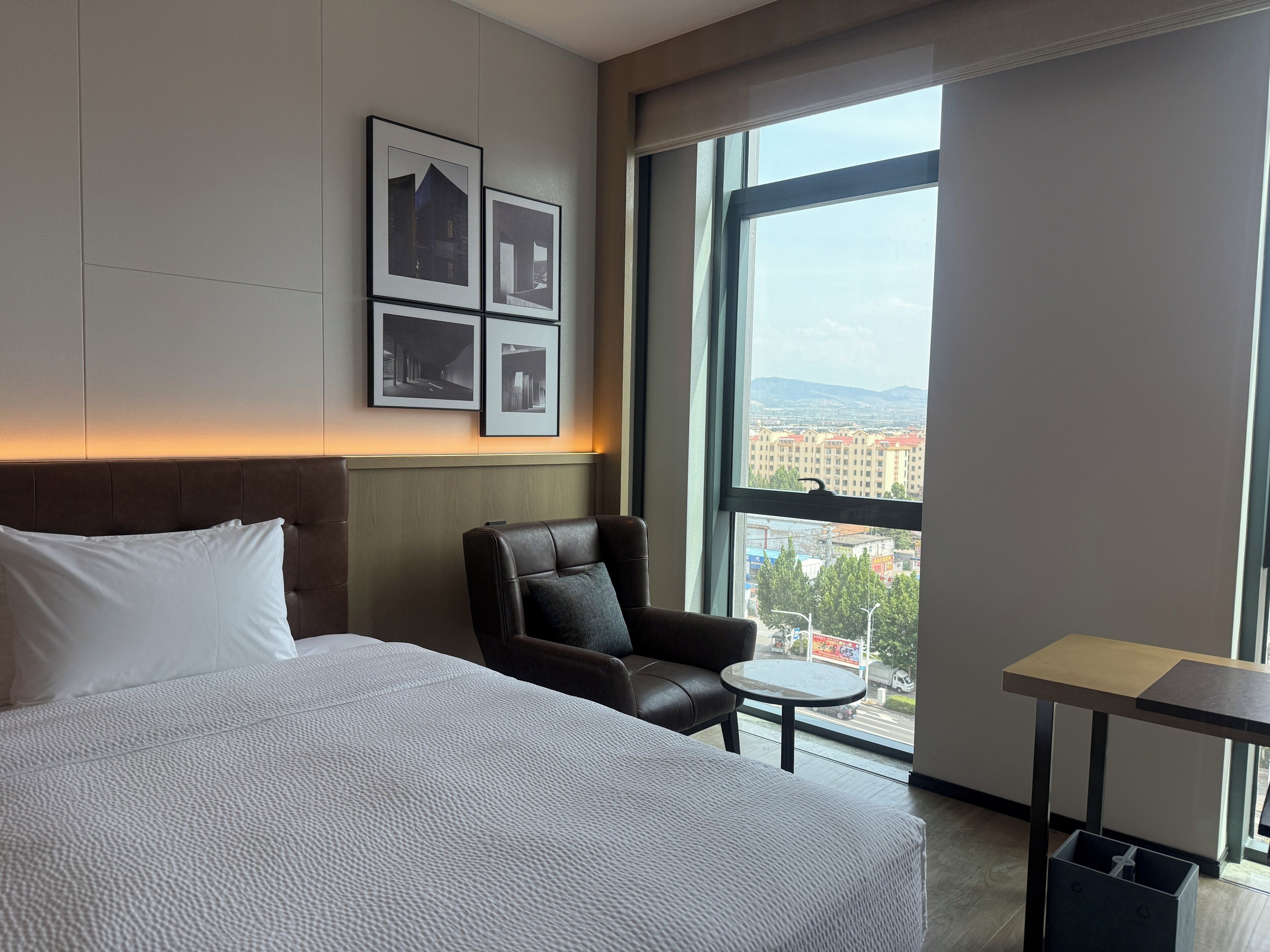 Habitación de hotel con una cama grande, un sillón de cuero, una mesita pequeña y vista a la ciudad a través de un ventanal amplio.