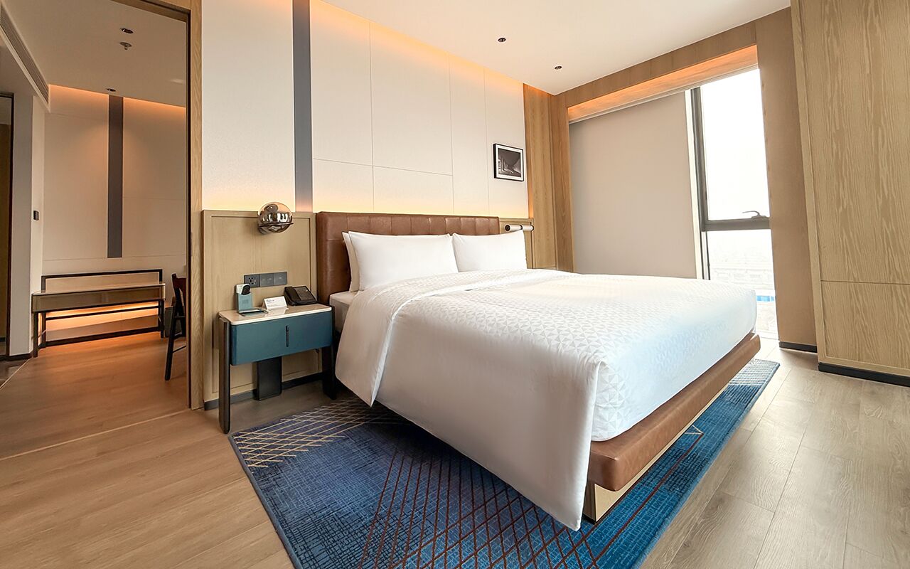 A hotel room with a large bed, a desk, a chair, a nightstand, and a window with a view.