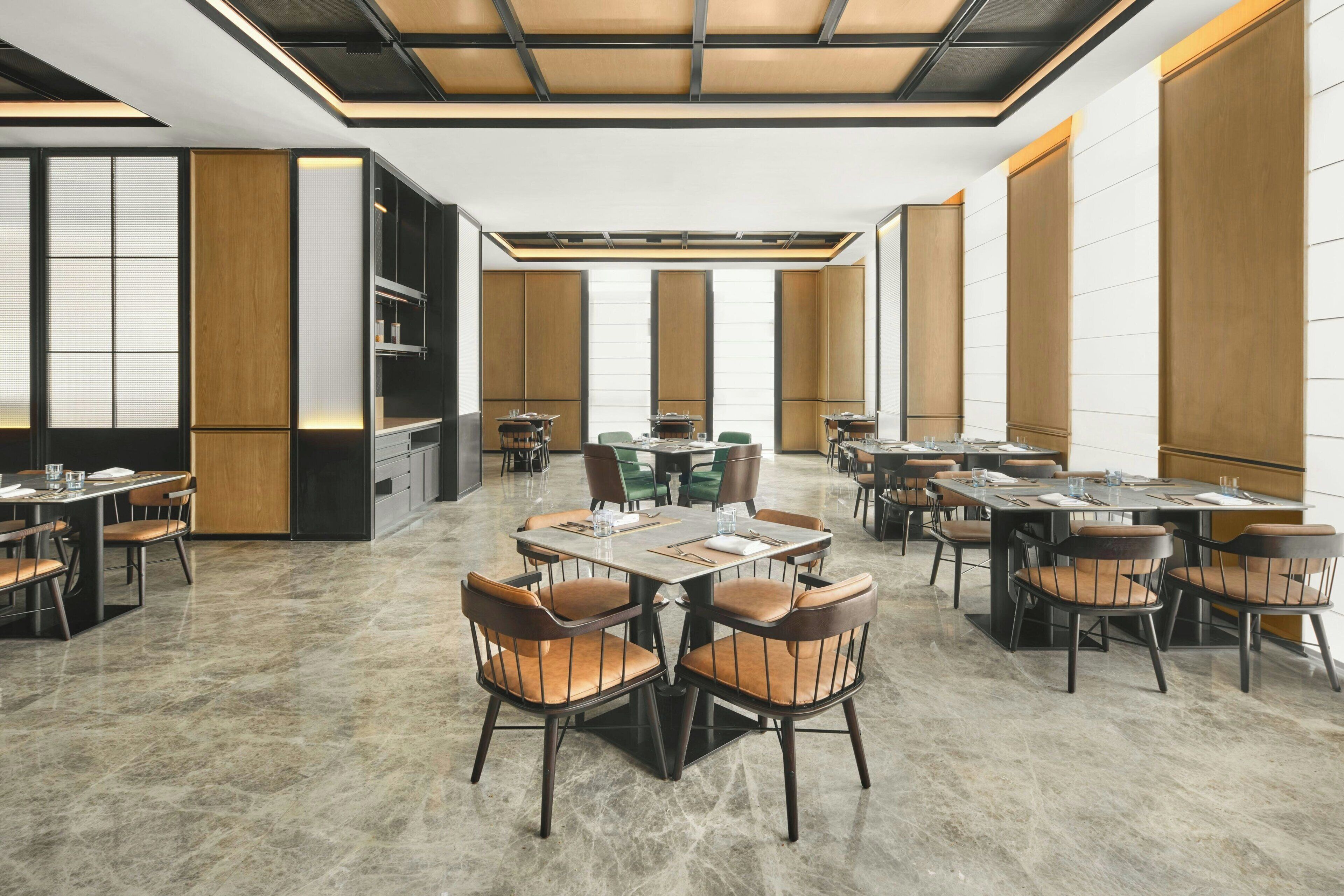 Un comedor de hotel espacioso con varias mesas y sillas, un diseño de techo moderno y grandes ventanales.