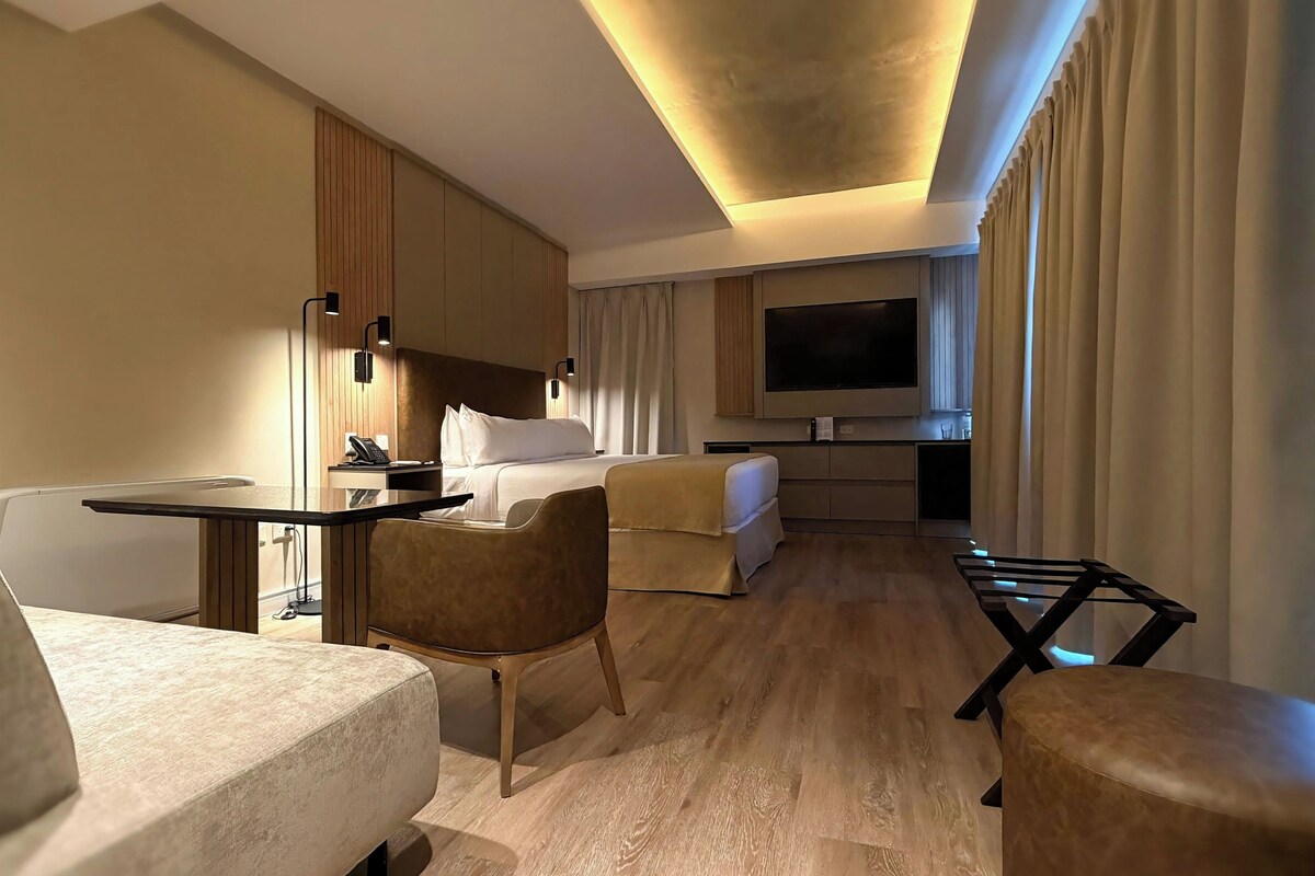 Ett hotellrum med en säng, ett skrivbord, en stol, en TV och en minibar.