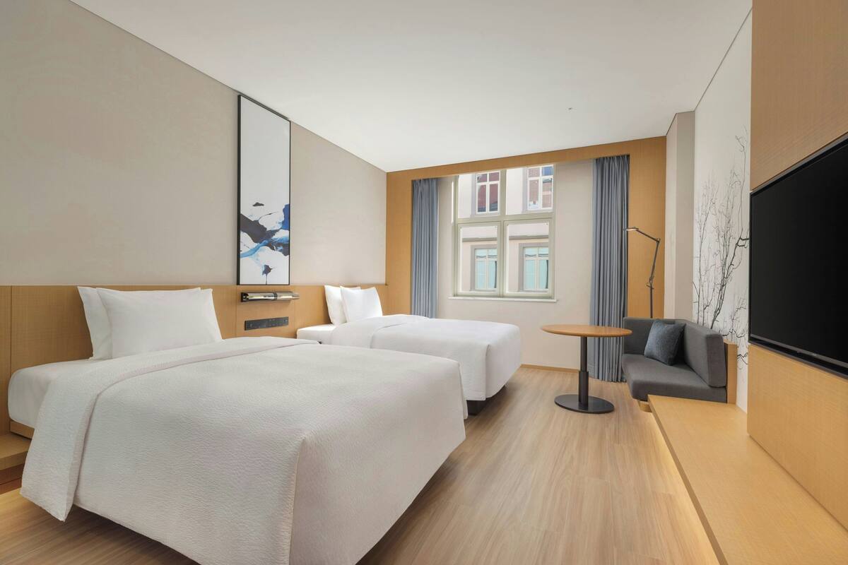 Habitación de hotel con dos camas, un sofá, un televisor, una mesita y una ventana con cortinas.