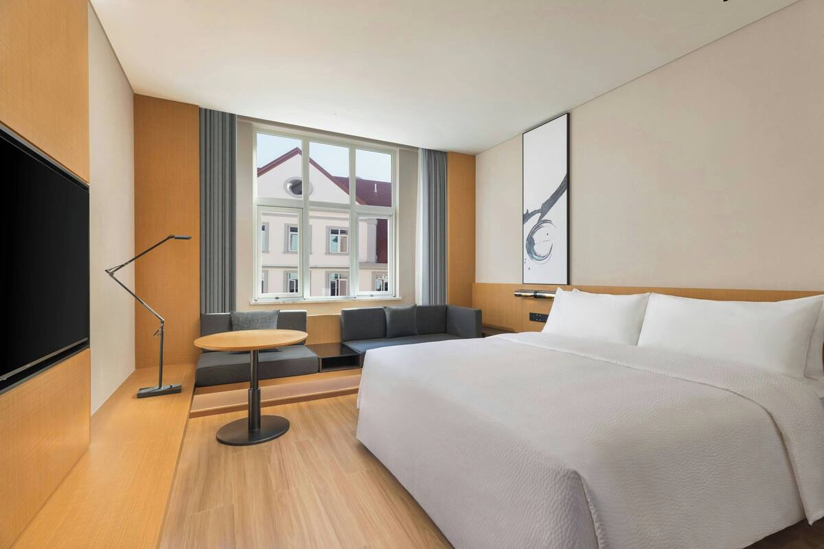 Habitación de hotel con cama, zona de estar, televisor y ventana con vistas a edificios.