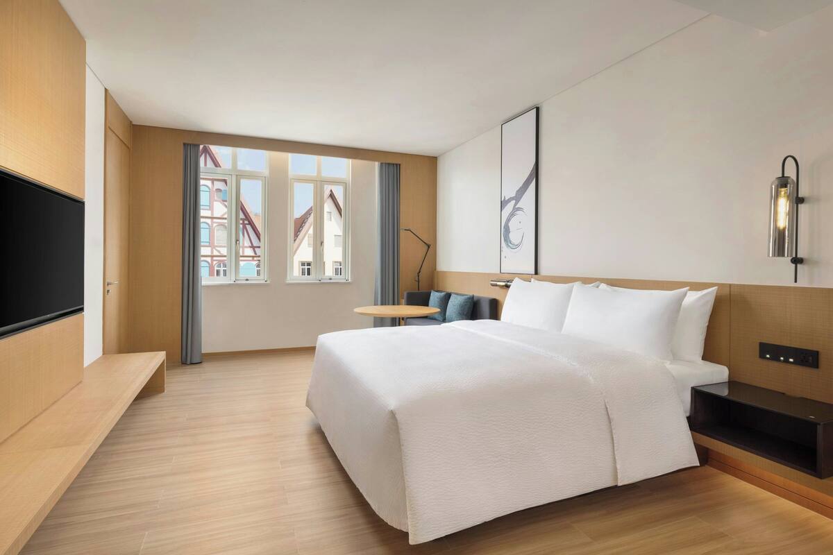 Una habitación de hotel moderna con una cama grande, un televisor de pantalla plana, una mesita pequeña y vistas a edificios a través de la ventana.