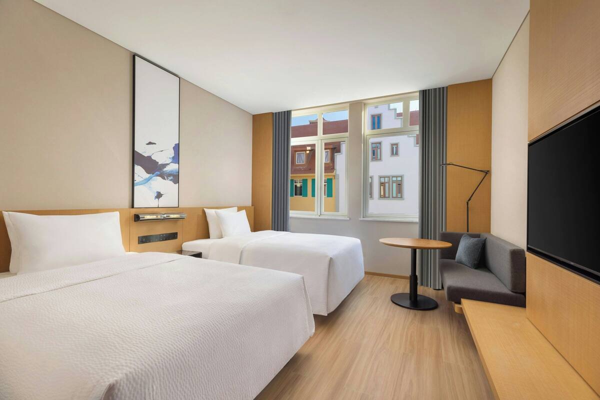 Habitación de hotel con dos camas, un sofá, una televisión, una mesita y una ventana con vistas a edificios.