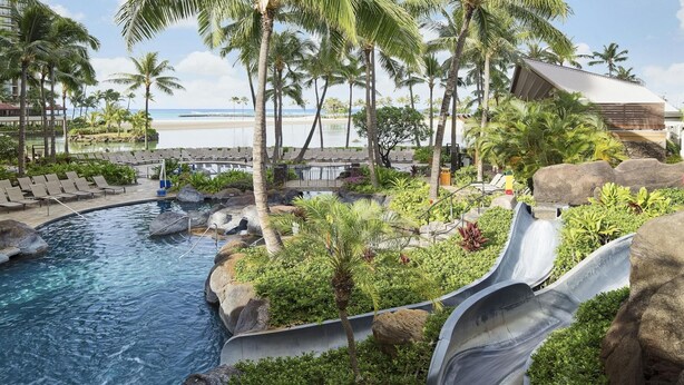 Ein Hotelpool mit Wasserrutsche, Liegestühlen, Palmen und Blick auf den Strand.
