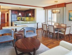 A spacious living area with a dining table, chairs, and a sofa. There is a kitchen in the background with wooden cabinets and a window. - Hawaiian Escape - Grand Waikikian- 2 Bedroom Plus with Partial Ocean View (Honolulu)