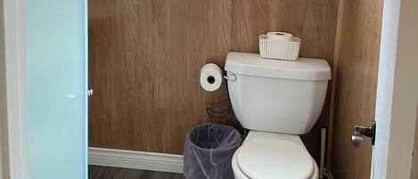 Et lille badeværelse med trævæg, et vindue, toilet, skraldespand og bruseområde.
