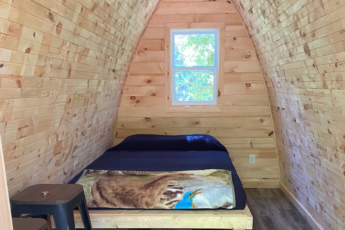 Pequeña habitación de ático con cama, ventana y dos taburetes.