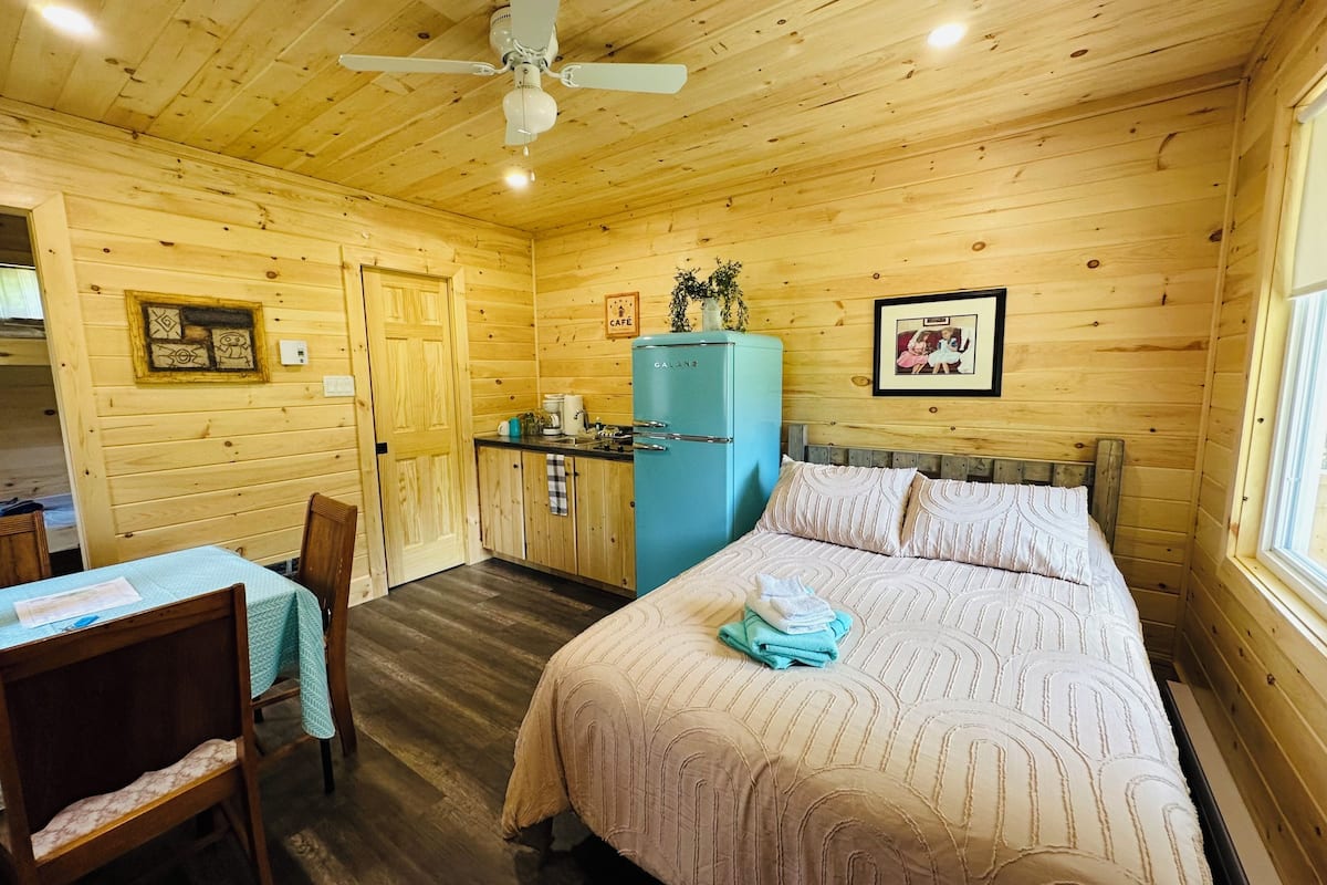 Interior de una cabaña de madera con cama, kitchenette, comedor y ventilador de techo.
