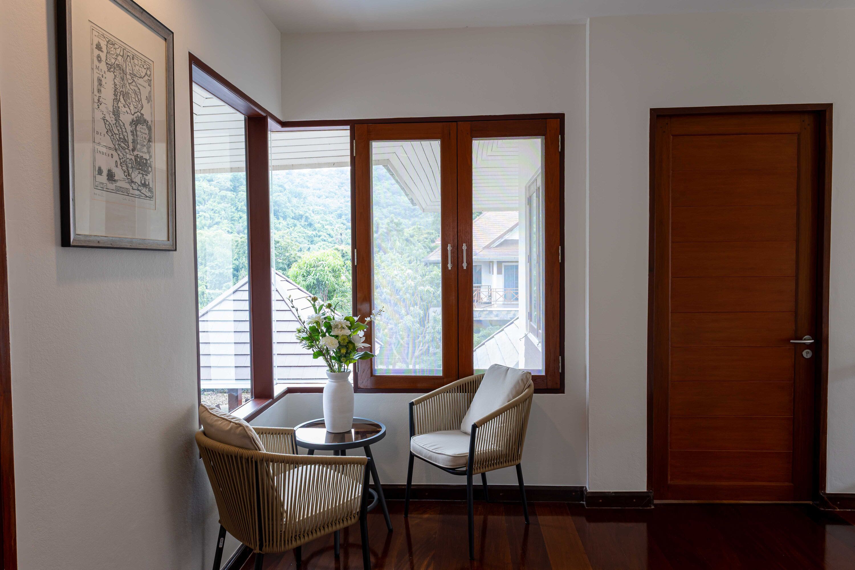 Una habitación con una puerta de madera, un mapa enmarcado en la pared, dos sillas de mimbre, una mesita redonda con un jarrón de flores y vistas a una casa a través de una ventana.
