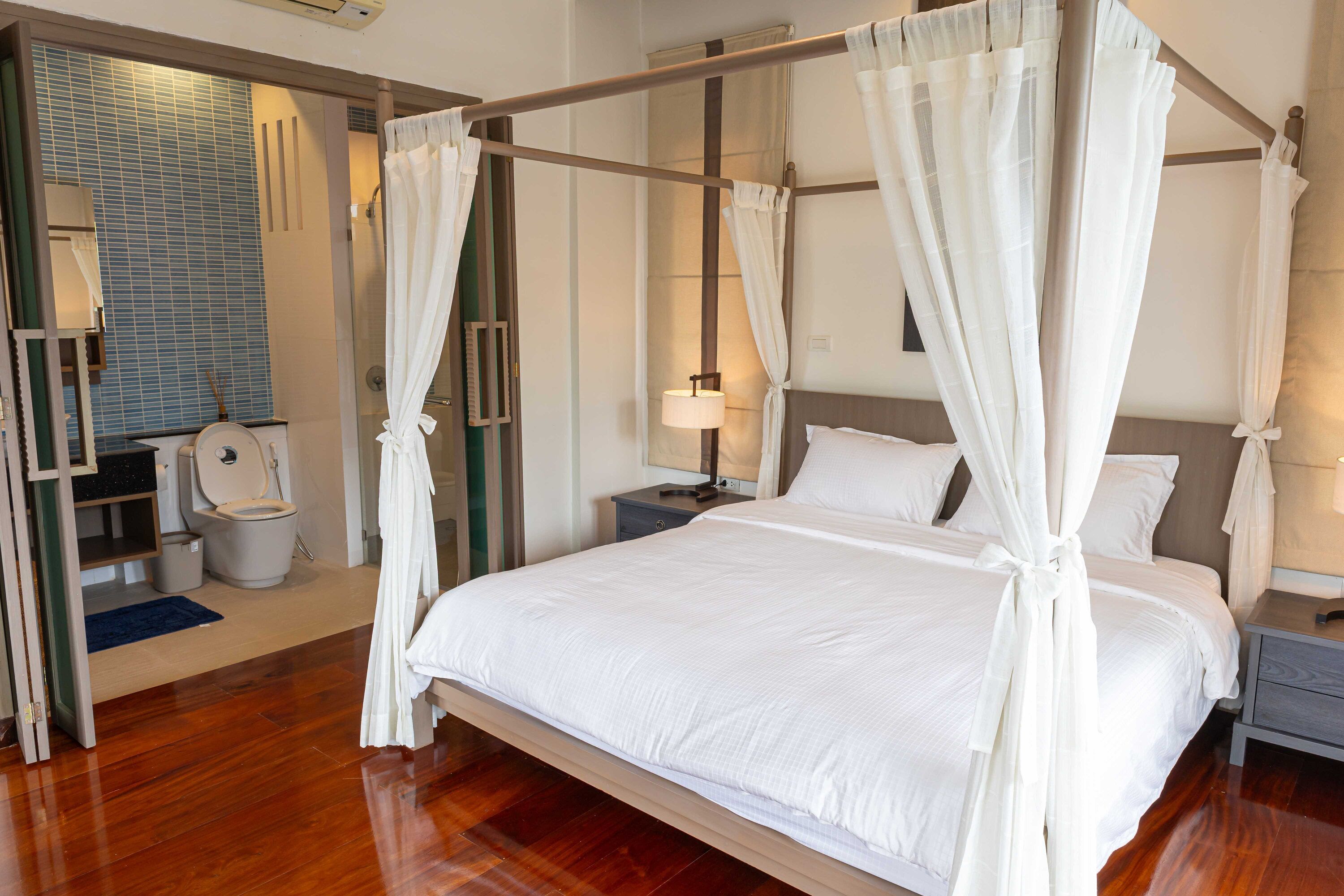 A four-poster bed with white linens, a wooden floor, a nightstand with a lamp, and a bathroom with a toilet and sink.