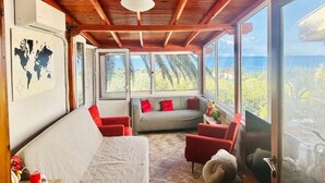 Une pièce chaleureuse avec un plafond en bois, un canapé, un fauteuil rouge et une grande fenêtre donnant sur l’extérieur.