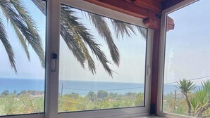 Une pièce avec un canapé gris et des coussins rouges, de grandes fenêtres offrant une vue sur les palmiers et l’océan.