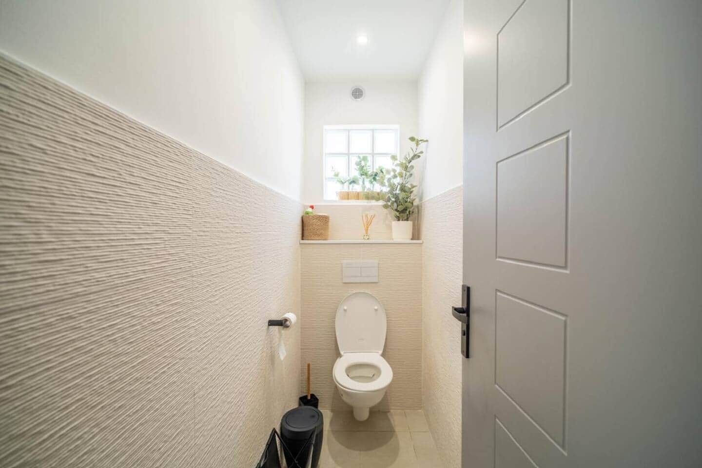 A narrow bathroom with a toilet, a trash bin, and a window with a plant.