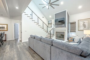 A spacious living area with a gray sectional sofa, a staircase, a flat-screen TV, and a fireplace. - Coastal Modern Oasis with Private Pool, Golf Cart, Putt-Putt | Steps to the Gulf (Panama City Beach)