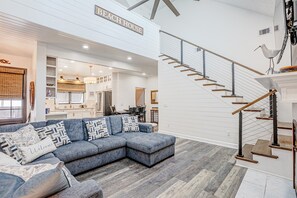 A modern living room with a sectional sofa, a staircase, and a kitchen area. - Coastal Modern Oasis with Private Pool, Golf Cart, Putt-Putt | Steps to the Gulf (Panama City Beach)