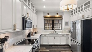 A modern kitchen with white cabinetry, stainless steel appliances, and a large window with blinds. - Coastal Modern Oasis with Private Pool, Golf Cart, Putt-Putt | Steps to the Gulf (Panama City Beach)