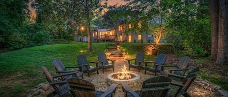 A fire pit surrounded by Adirondack chairs in a landscaped garden with a stone wall and a building in the background.
