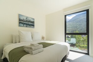 Une chambre à coucher avec un lit, des oreillers, une lampe de chevet, une fenêtre donnant sur la montagne et un tableau encadré au mur.