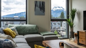 Una sala de estar moderna con un sofá seccional gris, una mesa de centro de madera y un ventanal con vistas a montañas y edificios.