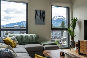 A modern living room with a gray sectional sofa, a wooden coffee table, and a large window offering a view of a mountain and buildings. - Barts Ski & Golf Retreat (Revelstoke)