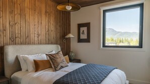 A bedroom with a wooden wall, a large window with a mountain view, a bed with white bedding and patterned pillows, and a floor lamp. - Max Send in Mackenzie Village (Revelstoke)