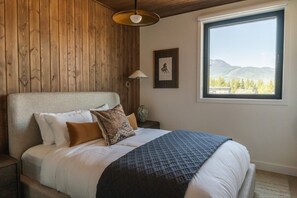 A bedroom with a wooden wall, a large window with a mountain view, a bed with white bedding and patterned pillows, and a floor lamp. - Max Send in Mackenzie Village (Revelstoke)