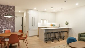 A modern kitchen with white cabinets, a central island with stools, and a dining area with a round table and orange chairs. - That 70s Chalet in the Mackenzie Village (Revelstoke)